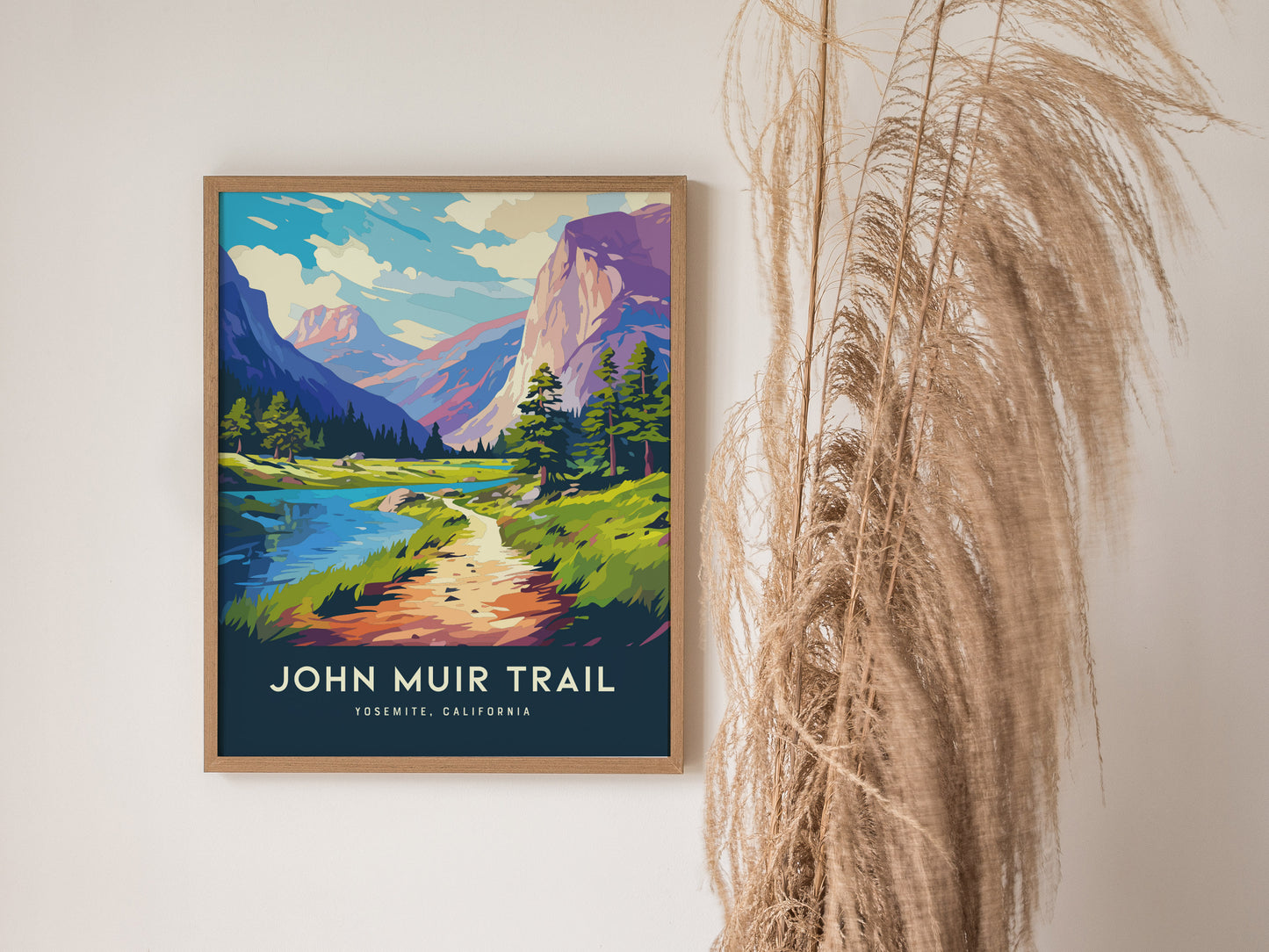 Framed John Muir Trail poster depicting a colorful landscape with mountains, trees, and a winding path. Tall dried grasses stand beside the artwork against a neutral wall.