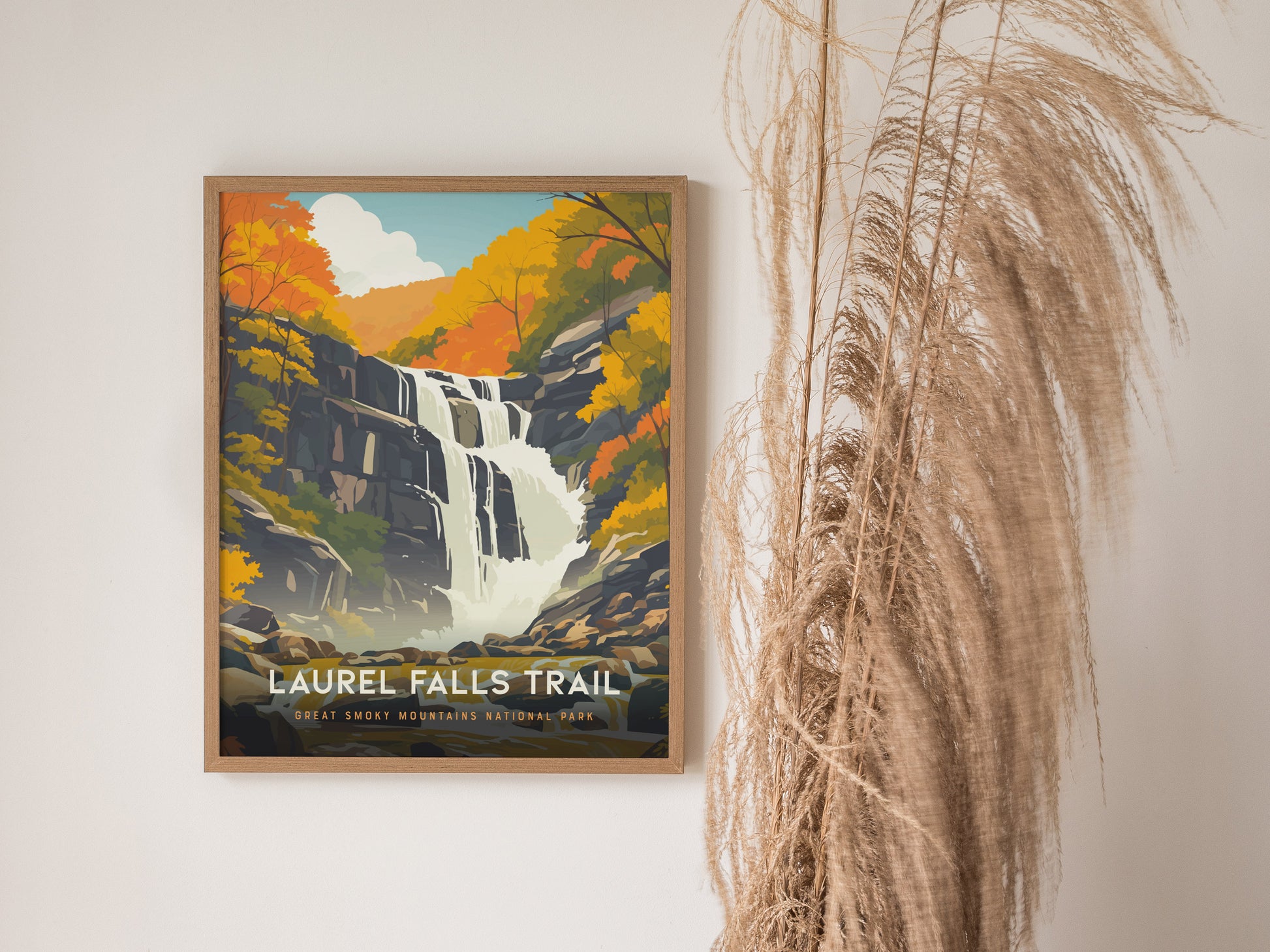 A framed poster of Laurel Falls Trail features a waterfall amid colorful autumn foliage. It hangs on a white wall beside tall, wispy dried grasses.
