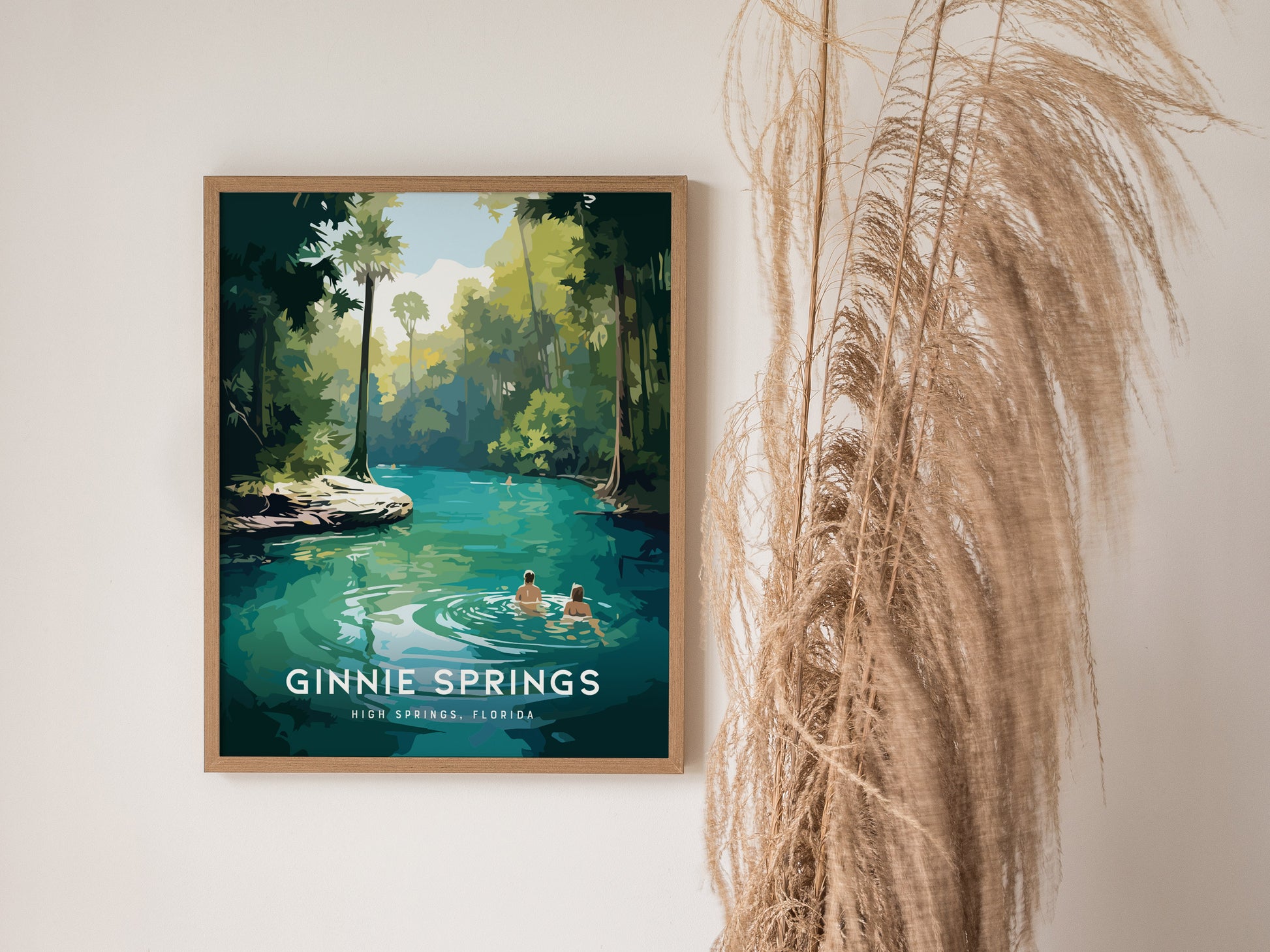 A framed poster of Ginnie Springs, Florida, depicts a serene turquoise water scene with two people swimming, surrounded by lush green foliage. Tall dried grasses are beside the artwork.