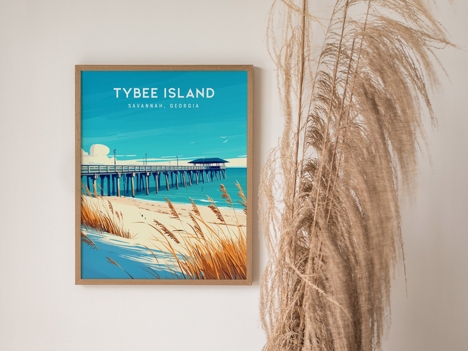 Framed artwork of Tybee Island features a pier extending into the ocean, with sandy dunes and sea oats in the foreground. Pampas grass decoratively placed beside the poster.