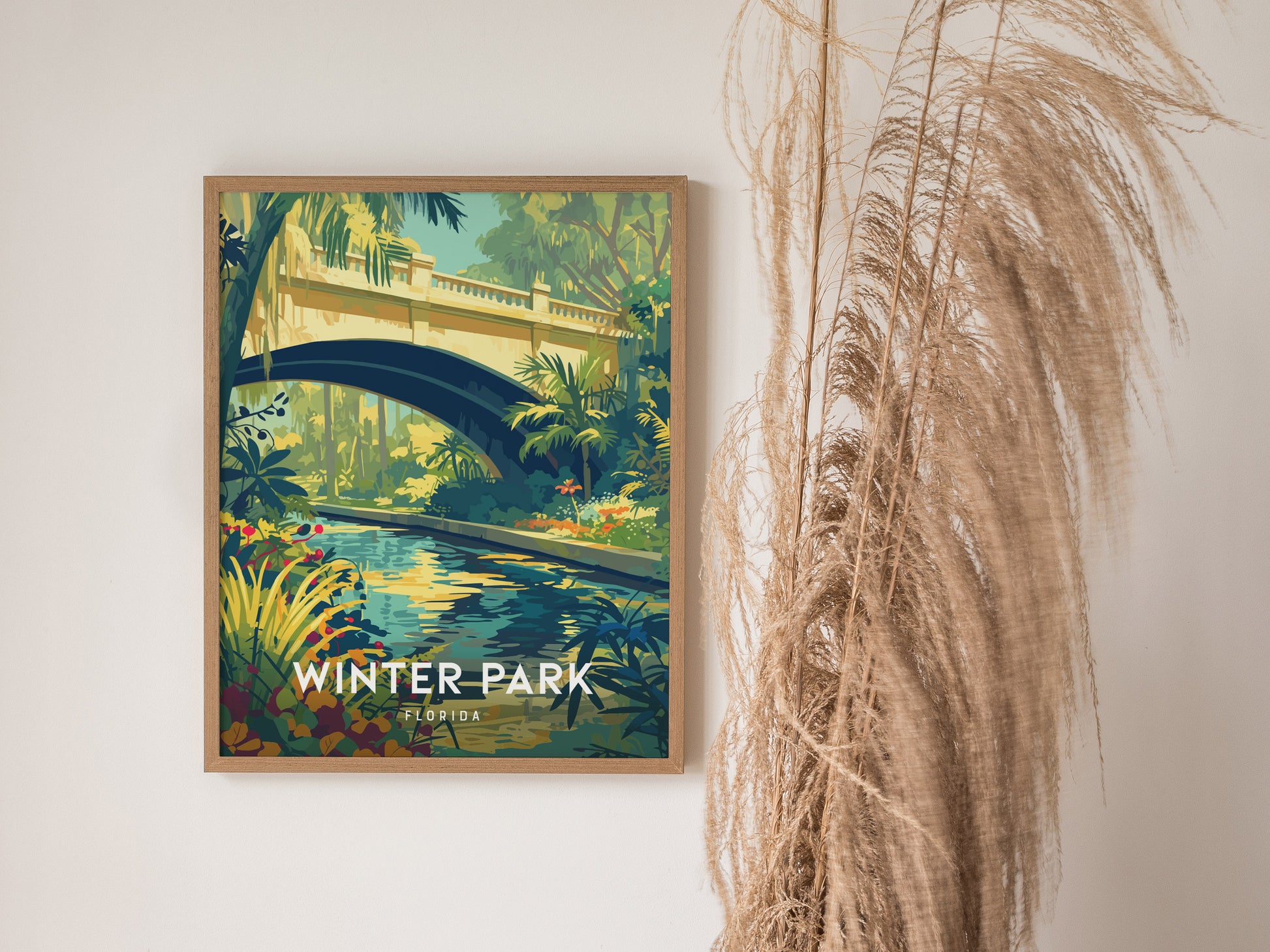 A framed poster of Winter Park, Florida, features a vibrant illustration of a lush garden with a stream flowing beneath an arched bridge. Tall grasses are positioned beside the artwork.
