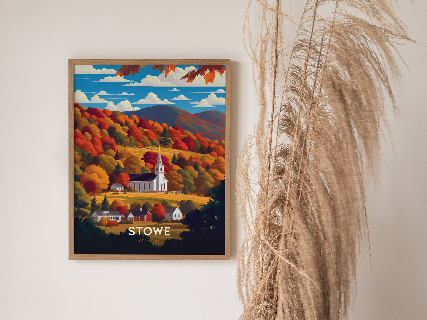 A framed poster depicting a white church steeple amidst vibrant autumn foliage in Stowe, Vermont, is displayed on a wall next to decorative pampas grass.