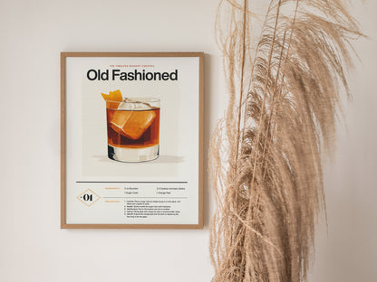 A framed Old Fashioned cocktail poster hangs on a white wall next to tall, dried pampas grass. The poster features an illustration of the drink and its recipe.