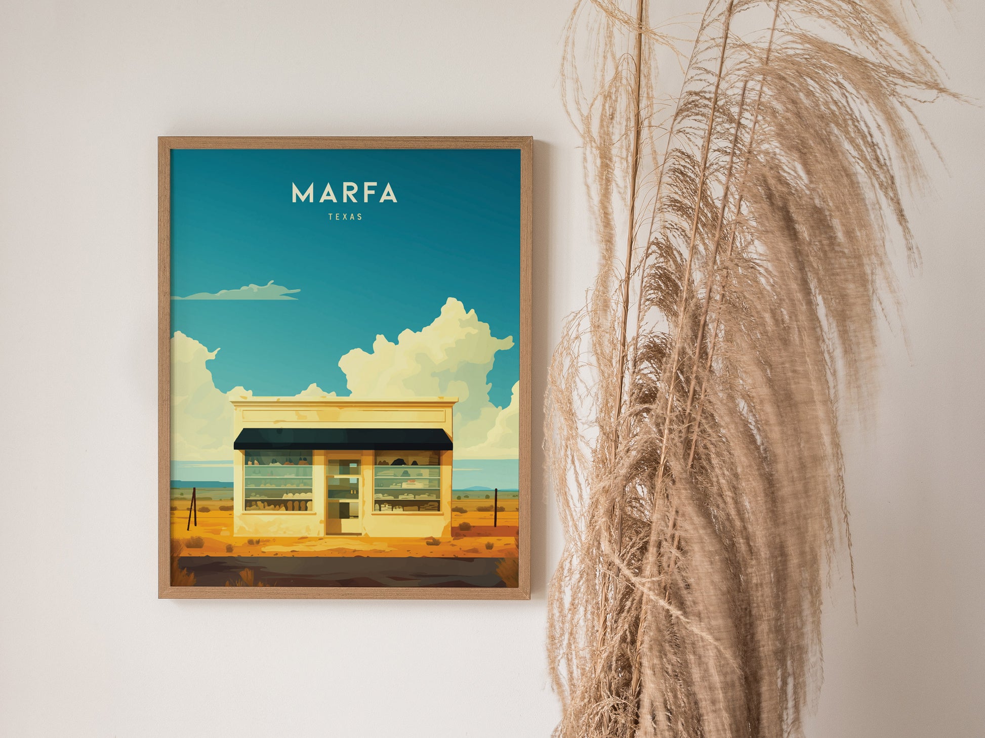 A framed poster of a minimalist building under a bright blue sky with clouds, labeled Marfa Texas, hangs on a wall next to tall, dried pampas grass.