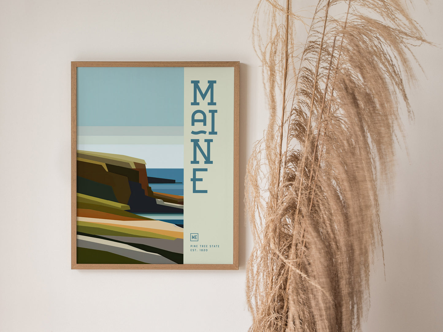 A framed minimalist poster featuring Maines coastline with abstract cliffs and ocean hues, accompanied by decorative text, is displayed next to tall, dried pampas grass.