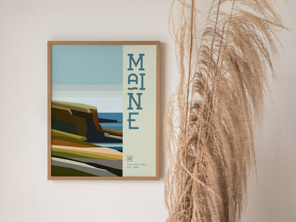 A framed minimalist poster featuring Maines coastline with abstract cliffs and ocean hues, accompanied by decorative text, is displayed next to tall, dried pampas grass.