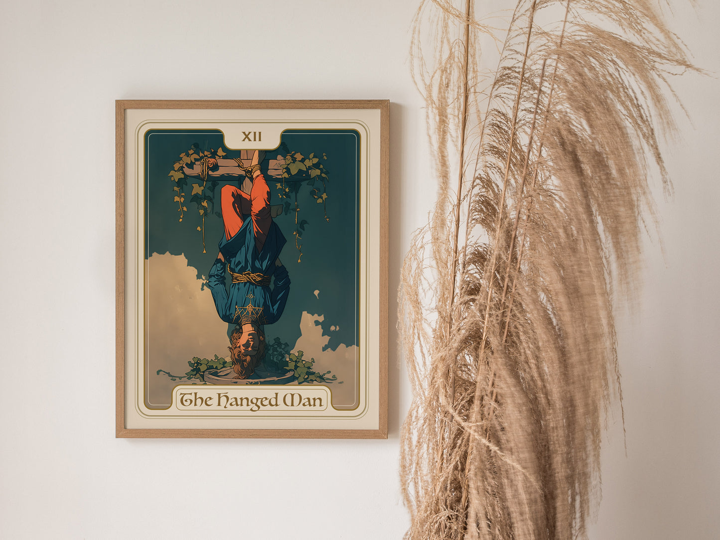 A framed poster of the Hanged Man Tarot Card XII is displayed on a wall, next to tall, dried pampas grass. The illustration depicts a serene figure suspended upside down.