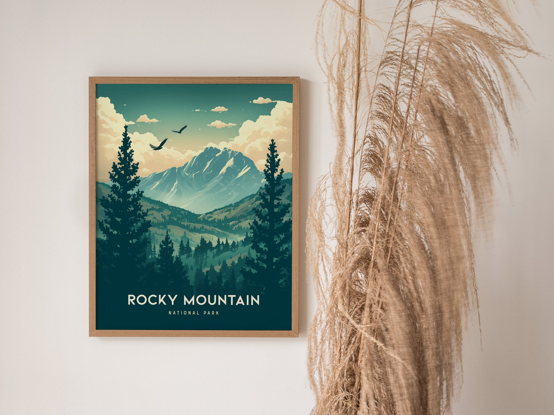 A framed Rocky Mountain National Park poster features snow-capped peaks and alpine forests. It hangs on a white wall beside tall, dried pampas grass.