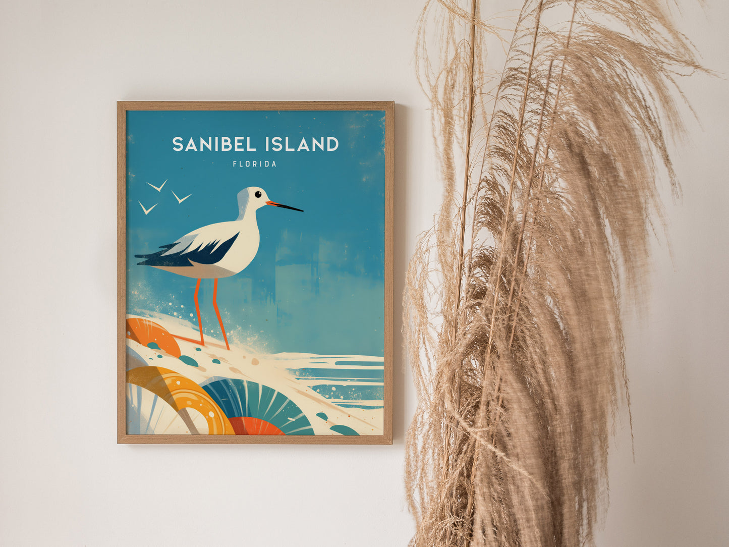A framed poster of Sanibel Island features an illustrated sandpiper on a beach with seashells. Decorative grasses are placed beside the artwork, enhancing the coastal theme.