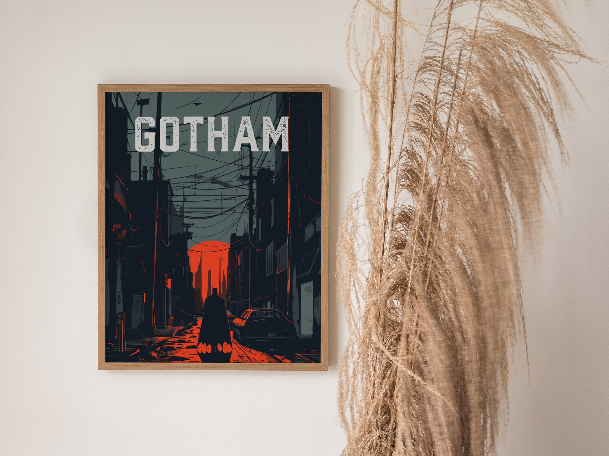 A framed poster titled Gotham depicts a dark cityscape in a noir style, with a shadowy figure standing in a dimly lit street under a red sky. The poster is displayed on a wall next to tall, dried pampas grass.