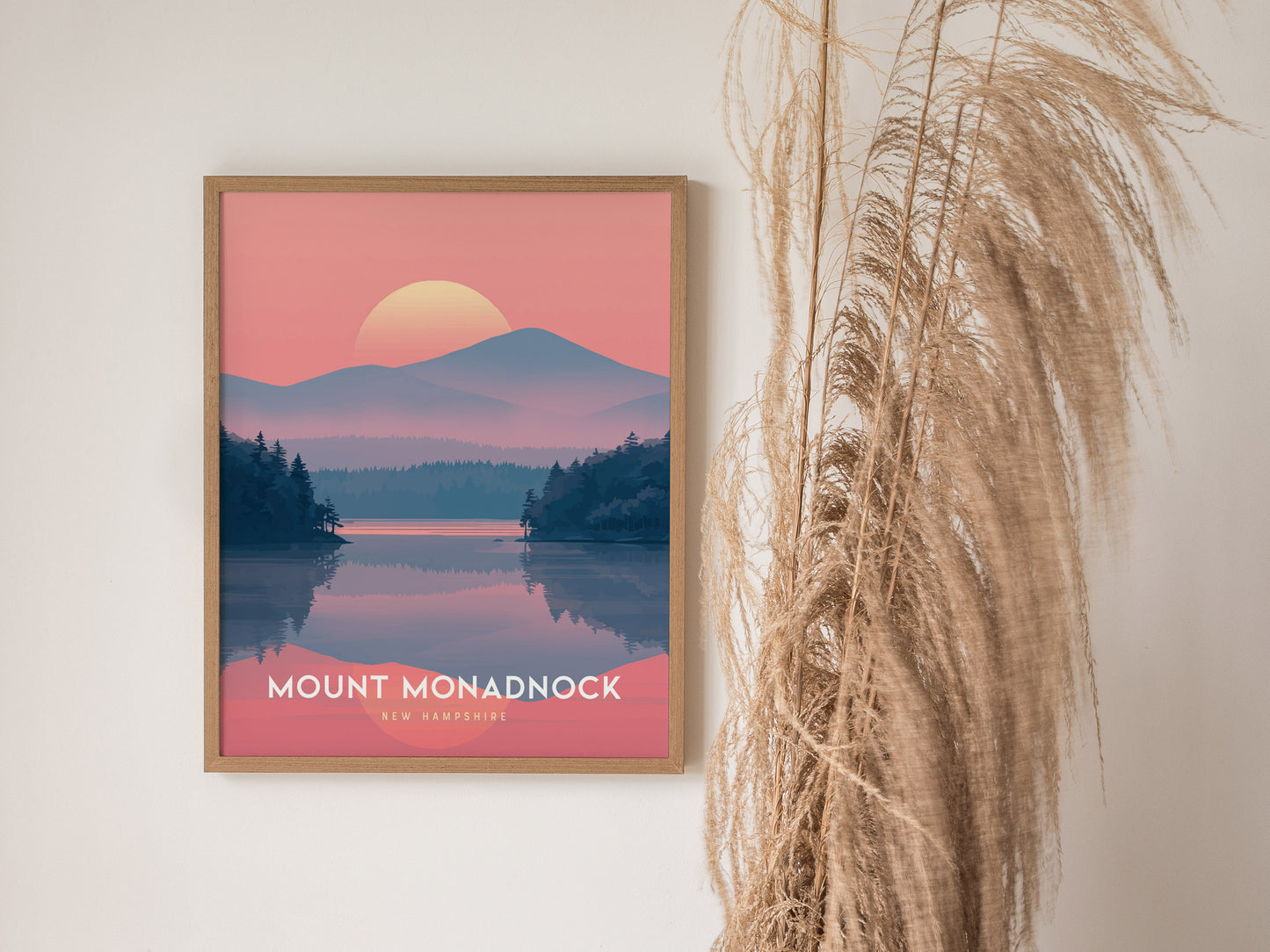 A framed Mount Monadnock poster features a stylized landscape with a sunset over mountains and a lake, set against a pale wall with dried pampas grass beside it.