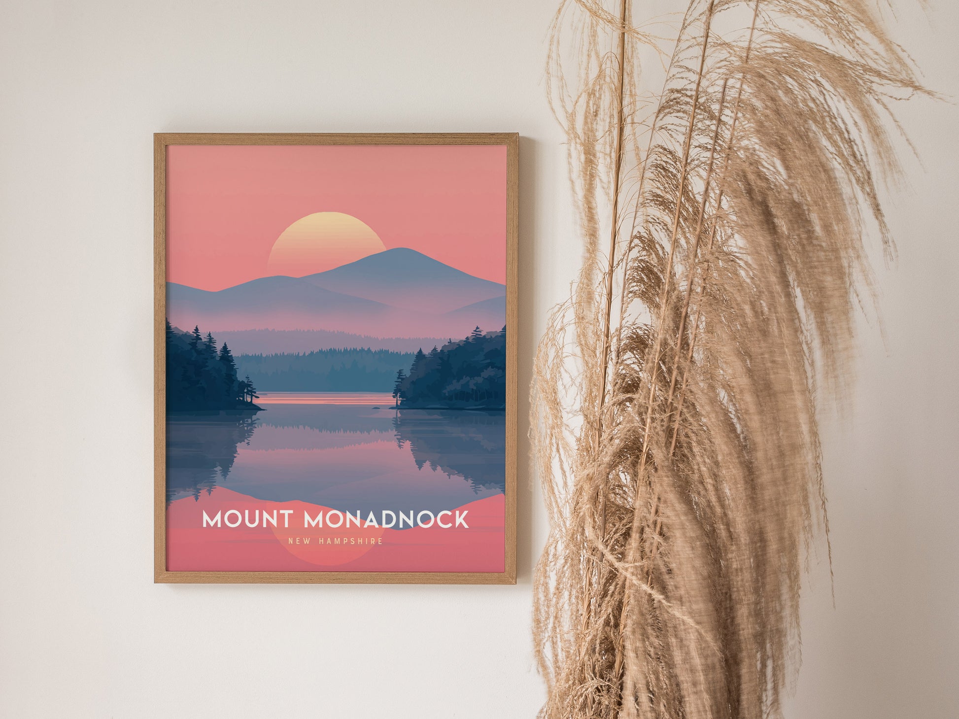 A framed Mount Monadnock poster features a stylized landscape with a sunset over mountains and a lake, set against a pale wall with dried pampas grass beside it.