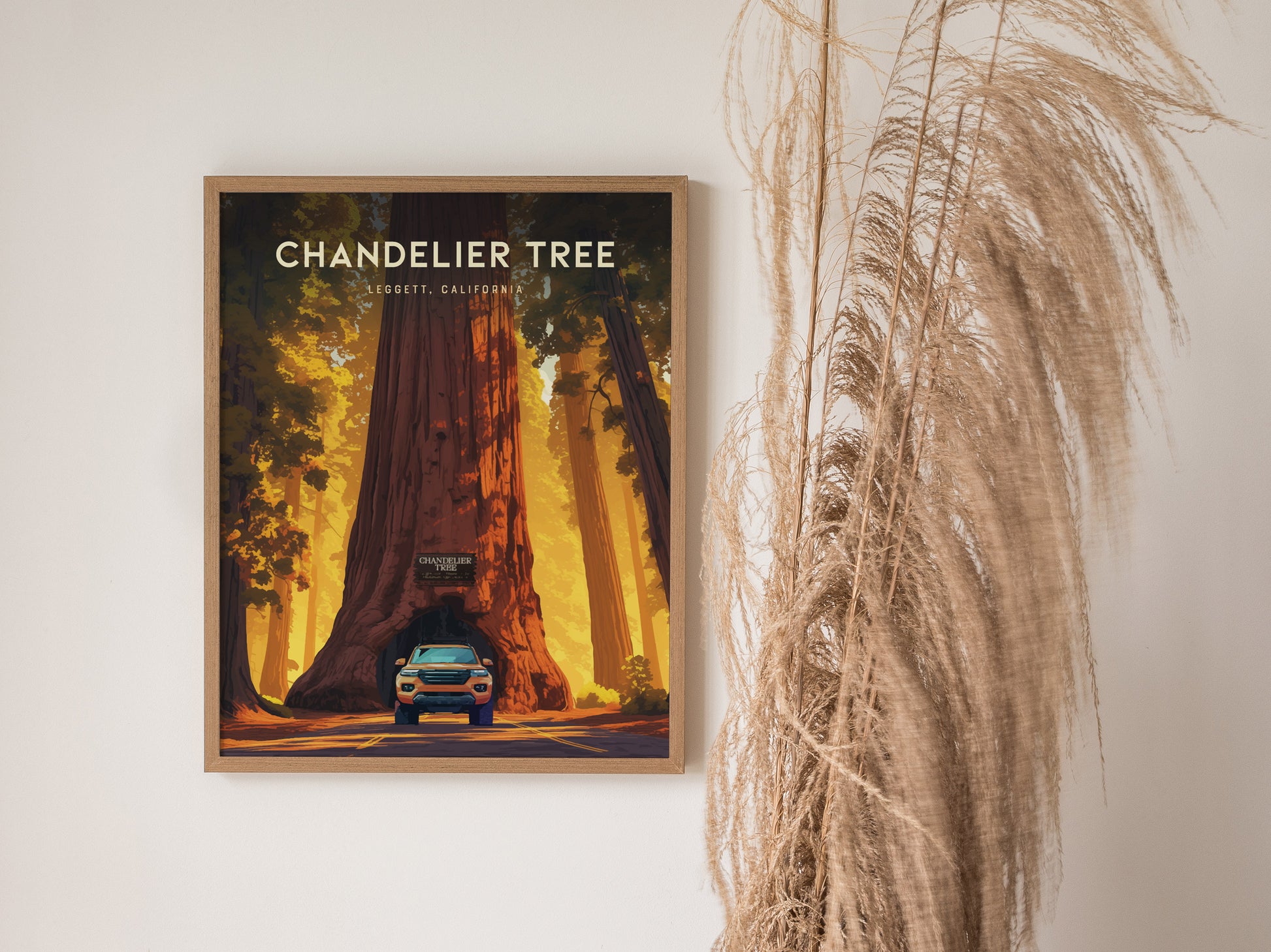 A framed poster of the Chandelier Tree in Leggett, California, featuring a car driving through its base, hangs on a wall next to decorative pampas grass.