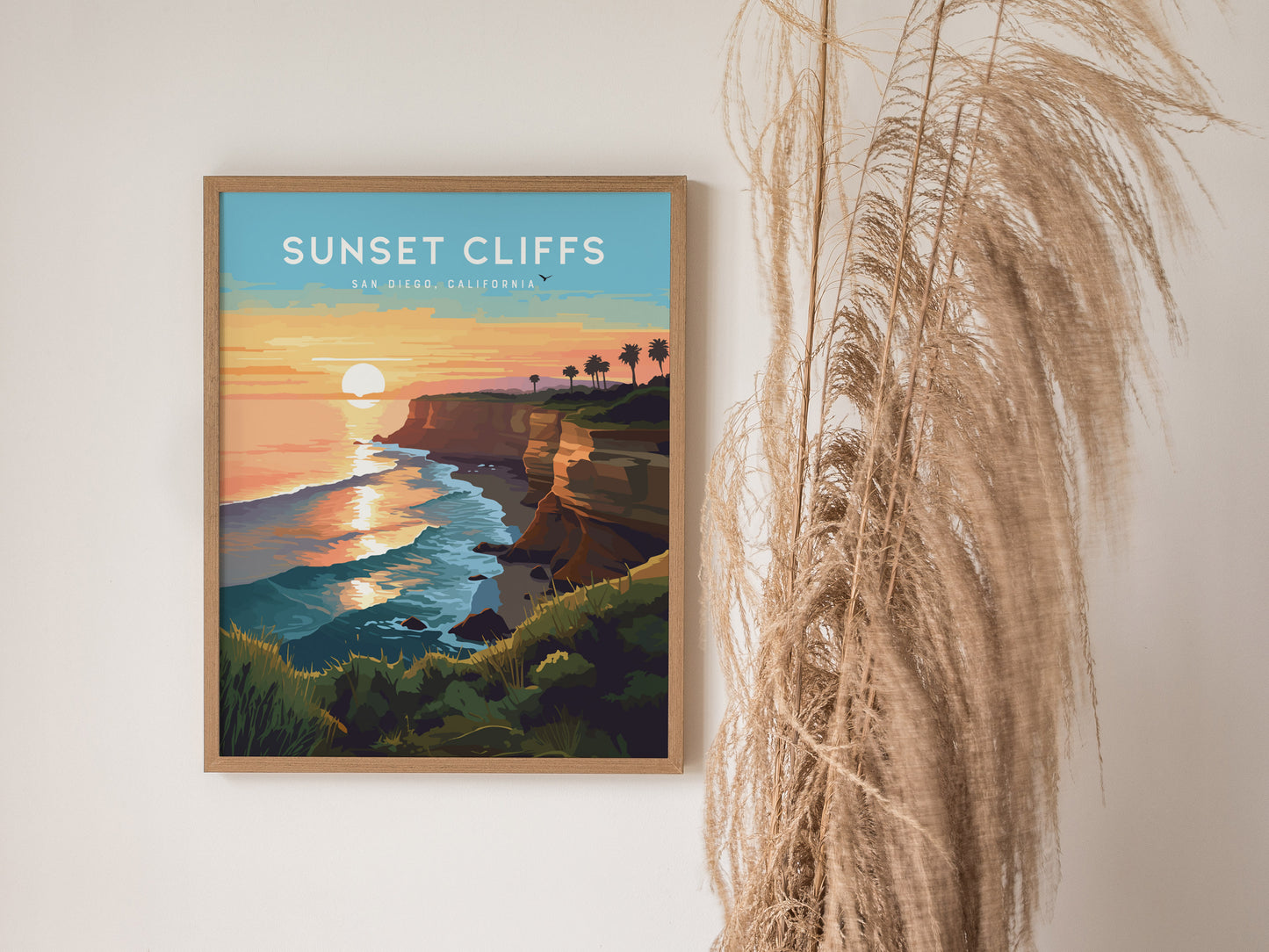 A framed poster of Sunset Cliffs in San Diego shows a vibrant sunset over the ocean, with cliffs and palm trees. Pampas grass decorates the right side of the scene.