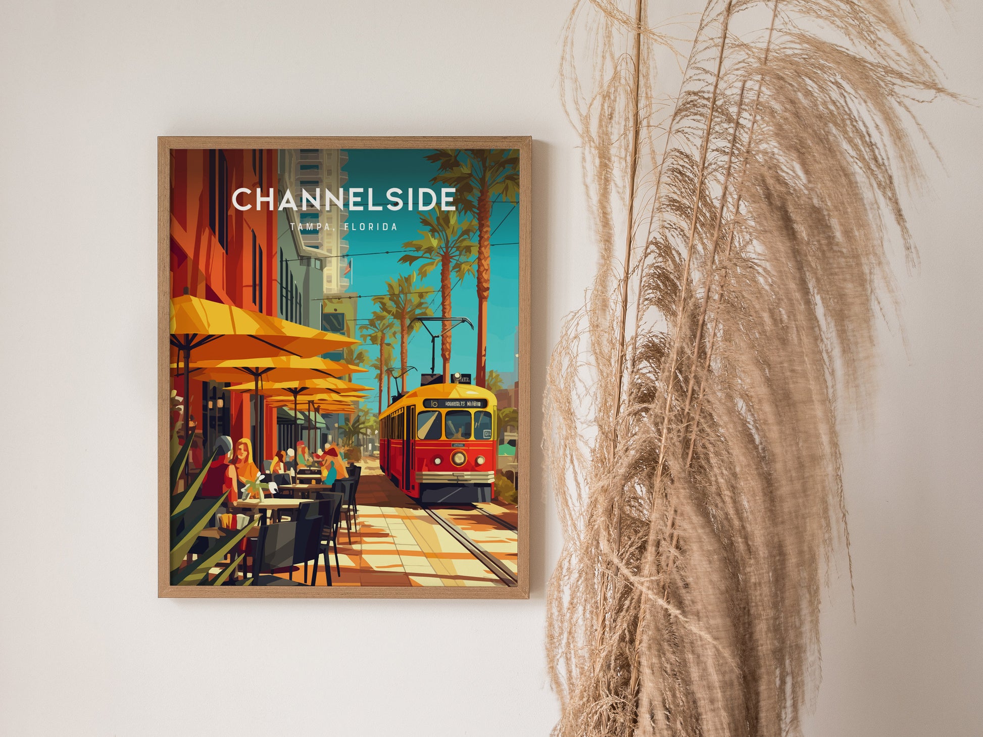 A framed Channelside Tampa poster features a vibrant street scene with a red trolley, yellow umbrellas, and palm trees, set against a white wall with decorative grass.