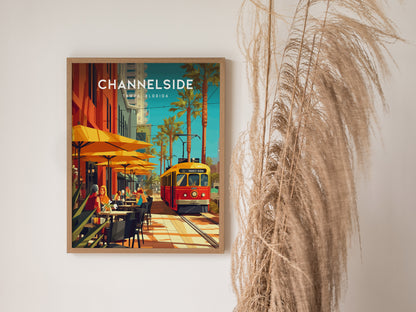 A framed Channelside Tampa poster features a vibrant street scene with a red trolley, yellow umbrellas, and palm trees, set against a white wall with decorative grass.
