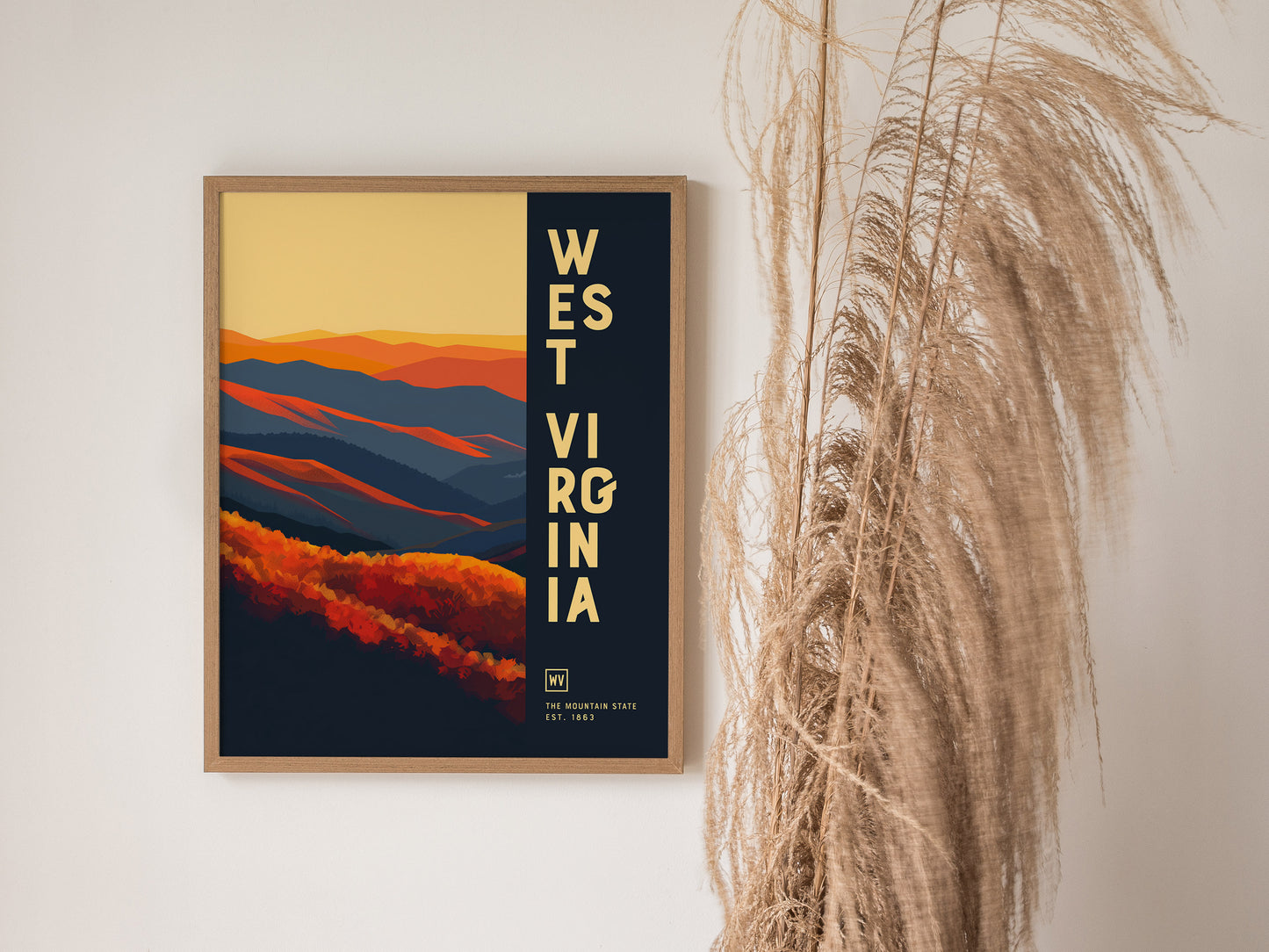 A framed poster of West Virginia features vibrant orange and blue abstract mountains against a yellow sky. It is displayed on a white wall next to tall dried grasses.