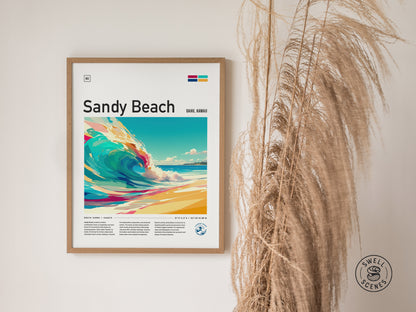 Framed Sandy Beach poster featuring vibrant, stylized waves and text, displayed on a wall beside dried pampas grass. The Swell Scenes logo is visible in the lower right corner.