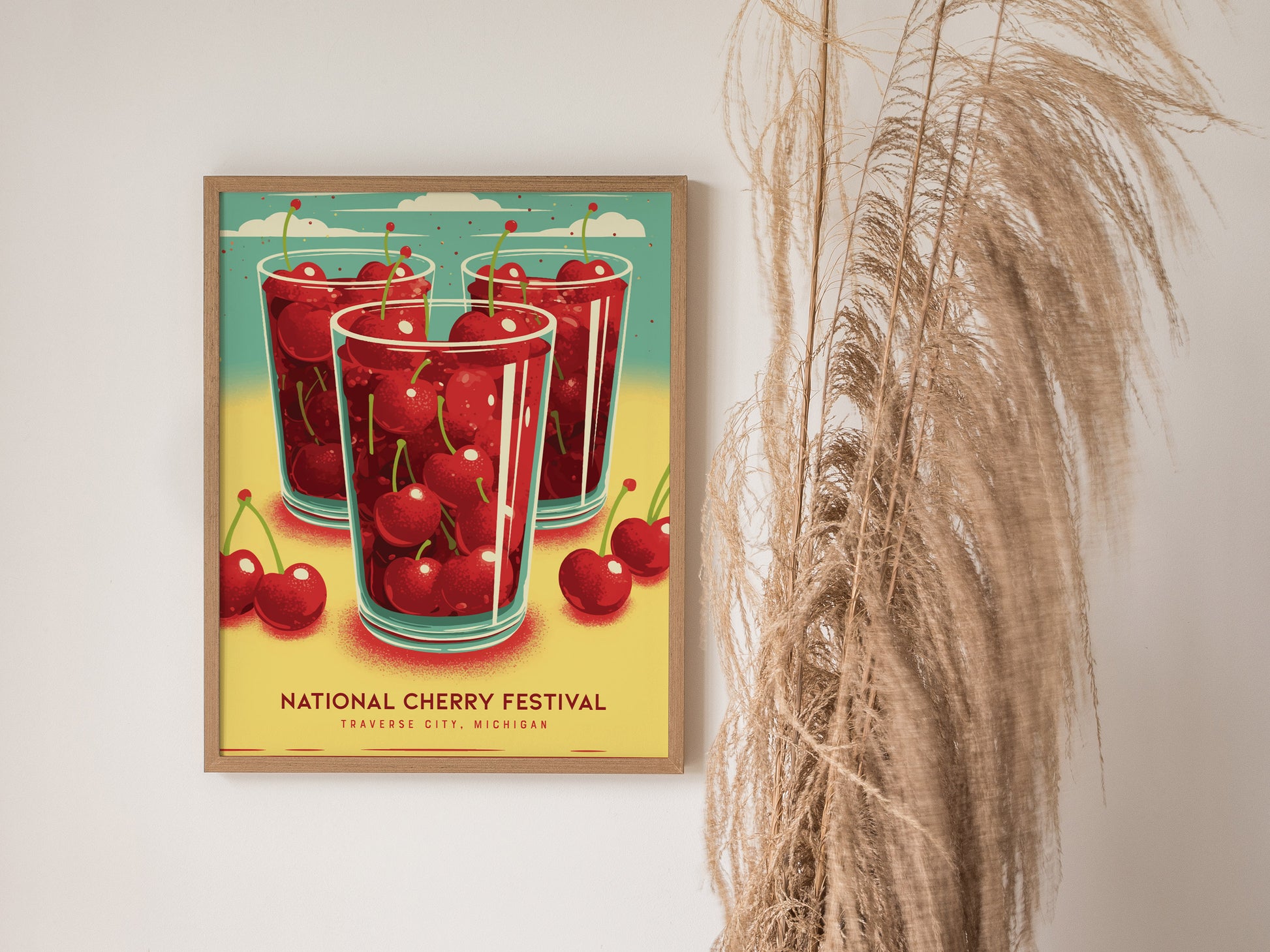 A framed poster on a wall features three glasses filled with cherries and the text National Cherry Festival, Traverse City, Michigan. Pampas grass decorates the right side.