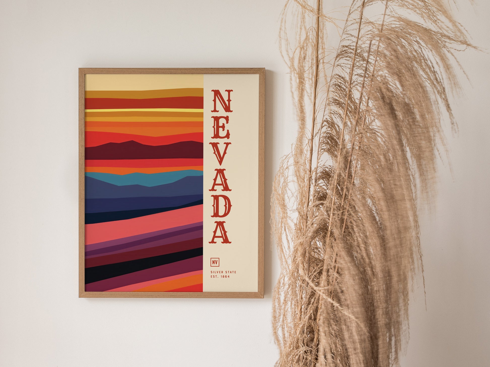 A framed Nevada poster with bold, colorful horizontal stripes and vertical text hangs on a white wall beside tall, dried pampas grass.