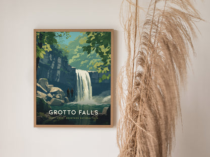 A framed poster of Grotto Falls hangs on a white wall, depicting a waterfall surrounded by lush greenery and rocks. Beside it, tall, dry pampas grass adds a natural decorative touch.