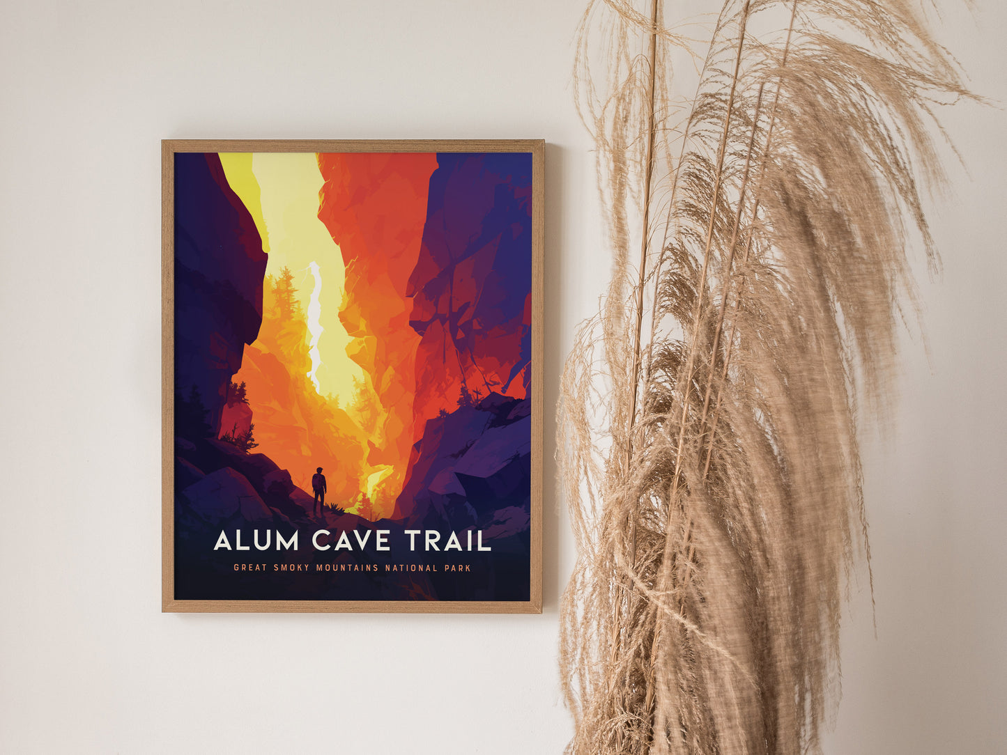 A framed poster titled Alum Cave Trail depicts a vibrant, colorful landscape with a lone hiker silhouetted against a dramatic canyon. Dried pampas grass decorates the scene.