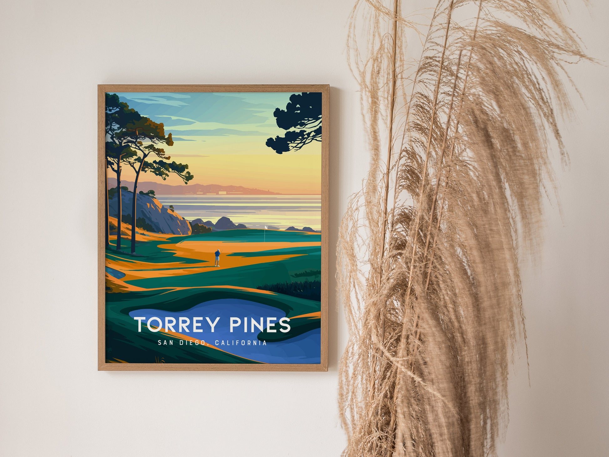 A framed poster of Torrey Pines Golf Course features vibrant coastal scenery with cliffs and a lone golfer, set against a sunset sky. Dried grasses are visible beside it.