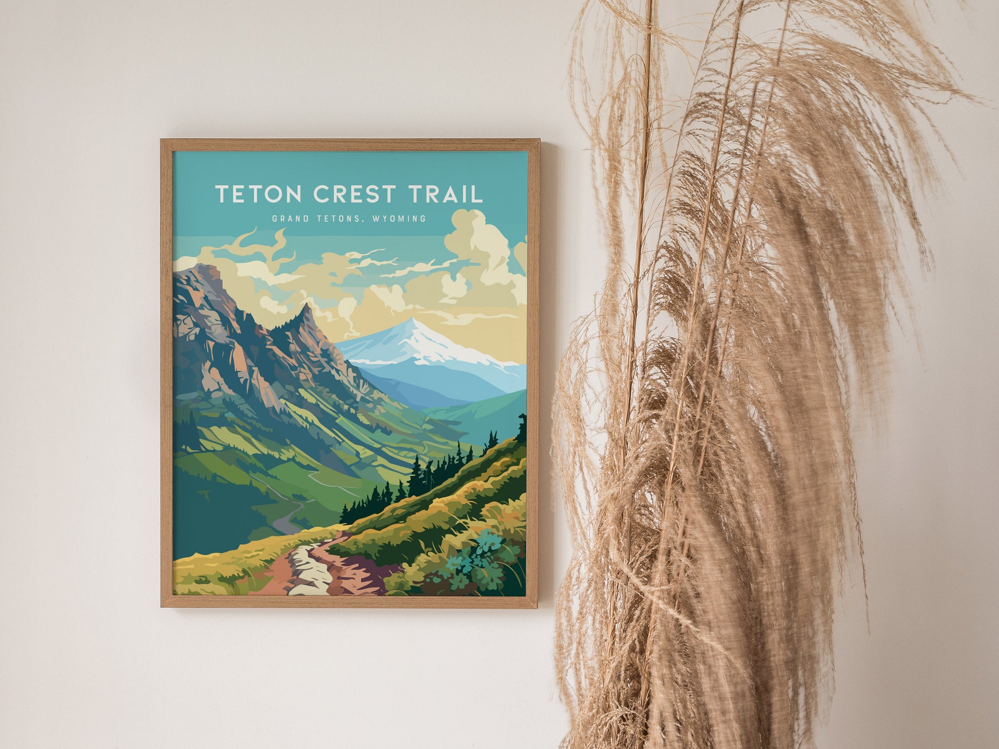 A framed poster of the Teton Crest Trail features vibrant mountains and a winding path under a blue sky, displayed on a wall beside dried decorative grasses.