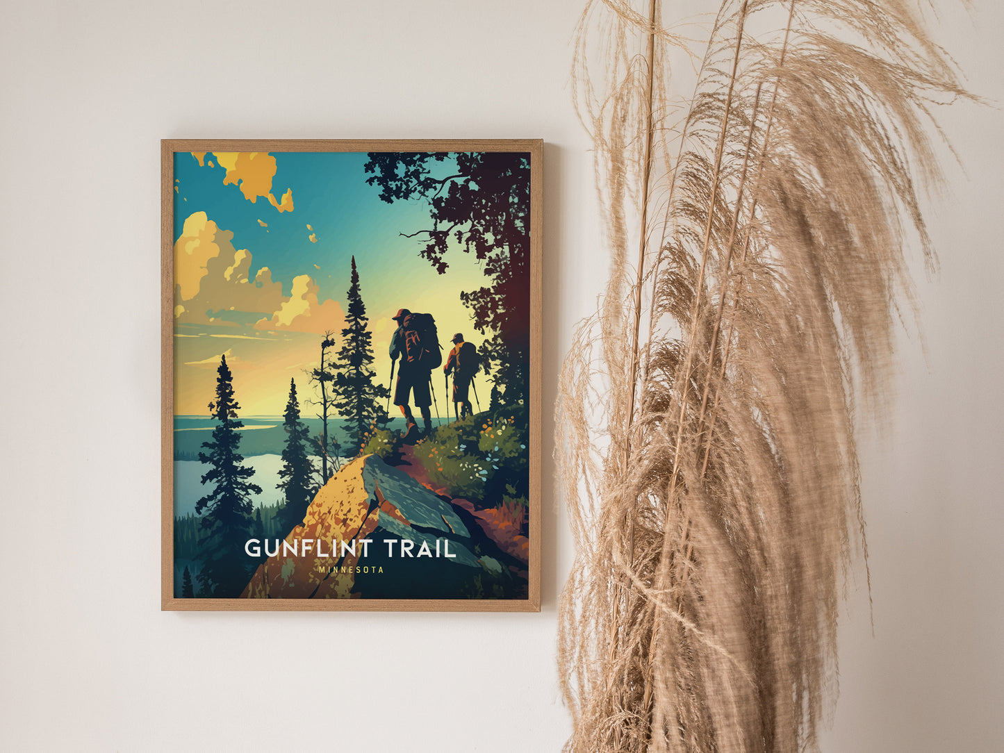 A framed poster titled Gunflint Trail depicts hikers on a scenic ridge surrounded by pine trees, with a dramatic sunset sky. Tall, dried grasses are positioned beside the artwork.
