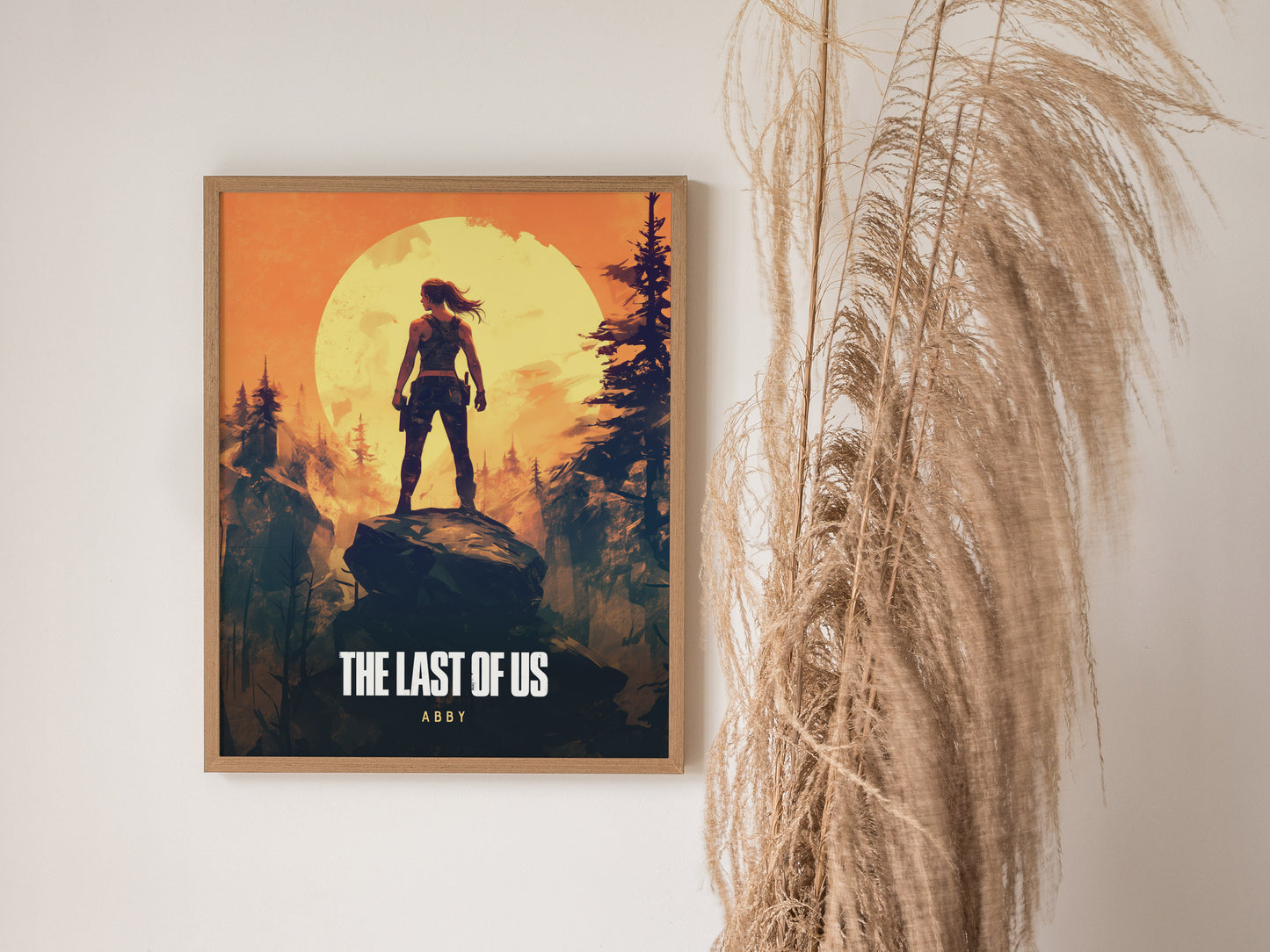 A framed poster of The Last of Us featuring a silhouetted figure standing on a rock against a bright orange sunset, next to dried pampas grass.