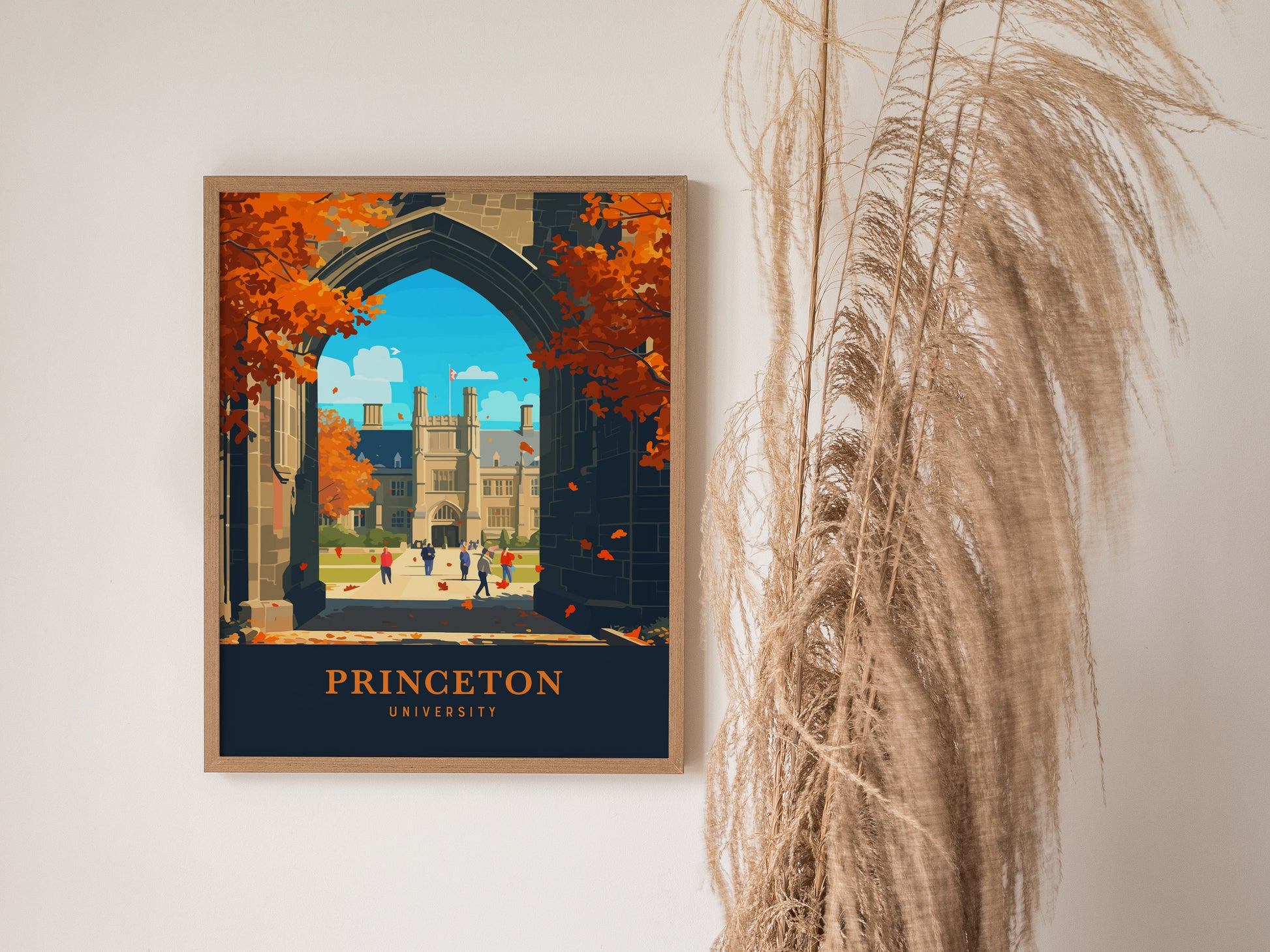 A framed poster of Princeton University featuring Gothic architecture and autumn trees is displayed on a white wall next to tall, wispy pampas grass.