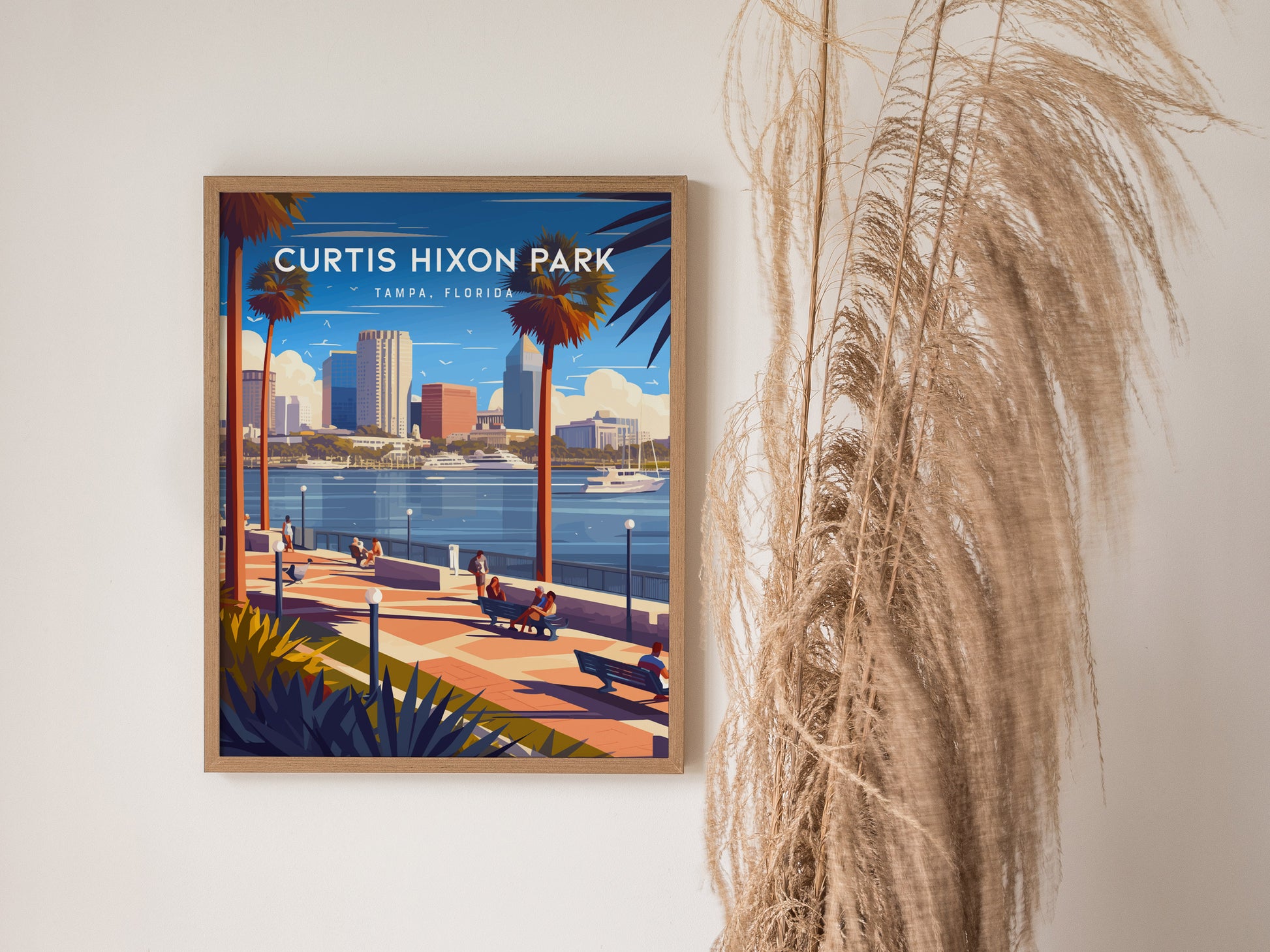 Framed poster of Curtis Hixon Park in Tampa, Florida, featuring a scenic view with palm trees, waterfront, skyscrapers, and people relaxing on benches. Pampas grass decorates the wall beside it.