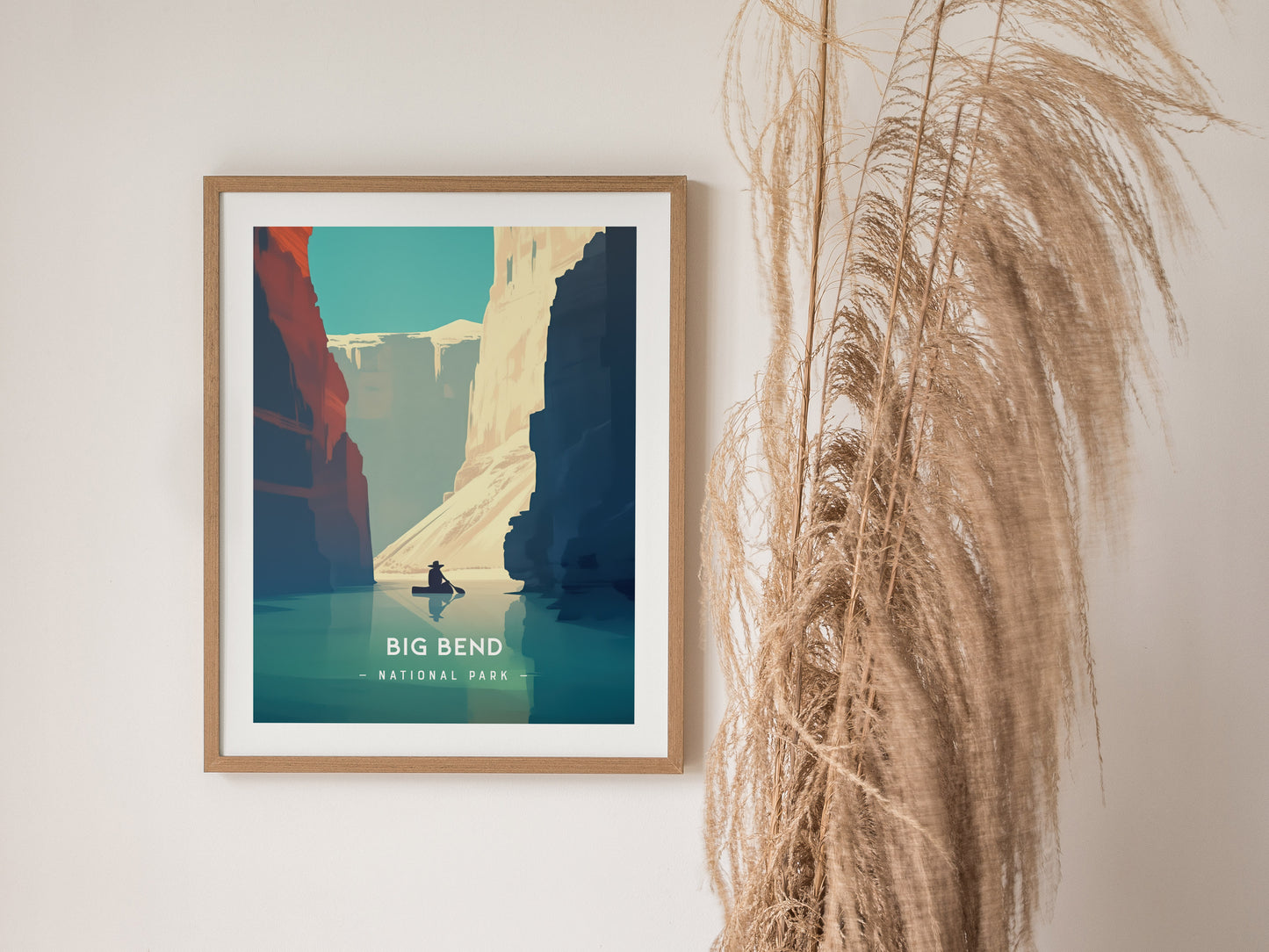 Framed Big Bend National Park poster featuring minimalist artwork of the Rio Grande canyon with a person kayaking. Tall dried grass decorates the wall beside the artwork.