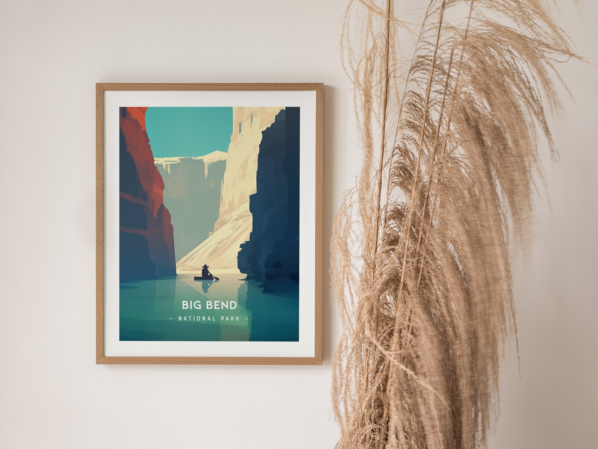 Framed Big Bend National Park poster featuring minimalist artwork of the Rio Grande canyon with a person kayaking. Tall dried grass decorates the wall beside the artwork.
