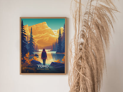 A framed Yoho National Park poster featuring a person standing by a tranquil lake, surrounded by evergreen trees and golden mountains under a vibrant sky. Pampas grass decorates the space.