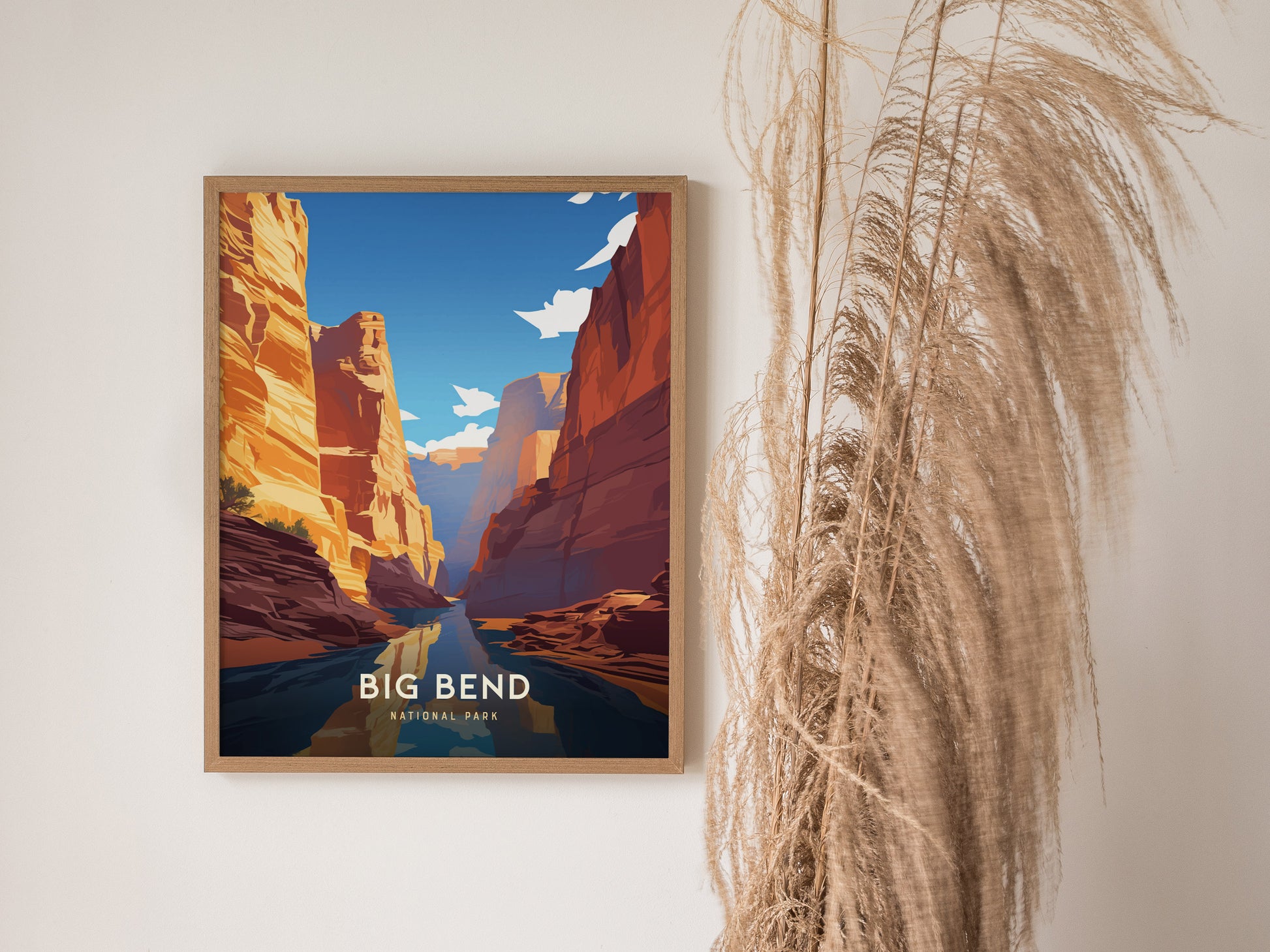 A framed poster of Big Bend National Park hangs on a white wall, featuring vibrant canyon landscapes and a clear blue sky, with dried pampas grass nearby for decoration.