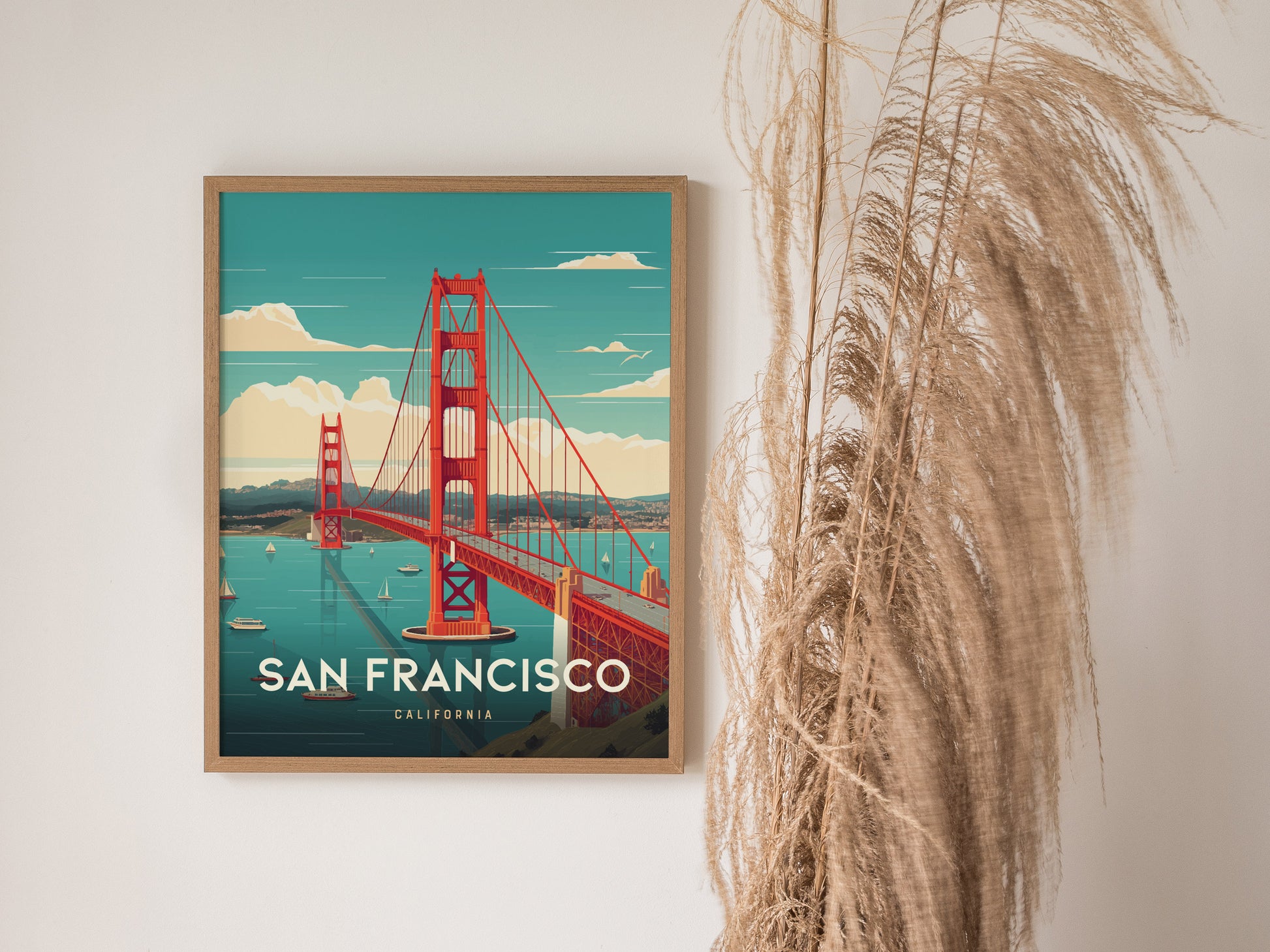 A framed poster of the Golden Gate Bridge with a turquoise sky and sailboats in the bay, displayed next to tall, dried ornamental grass.
