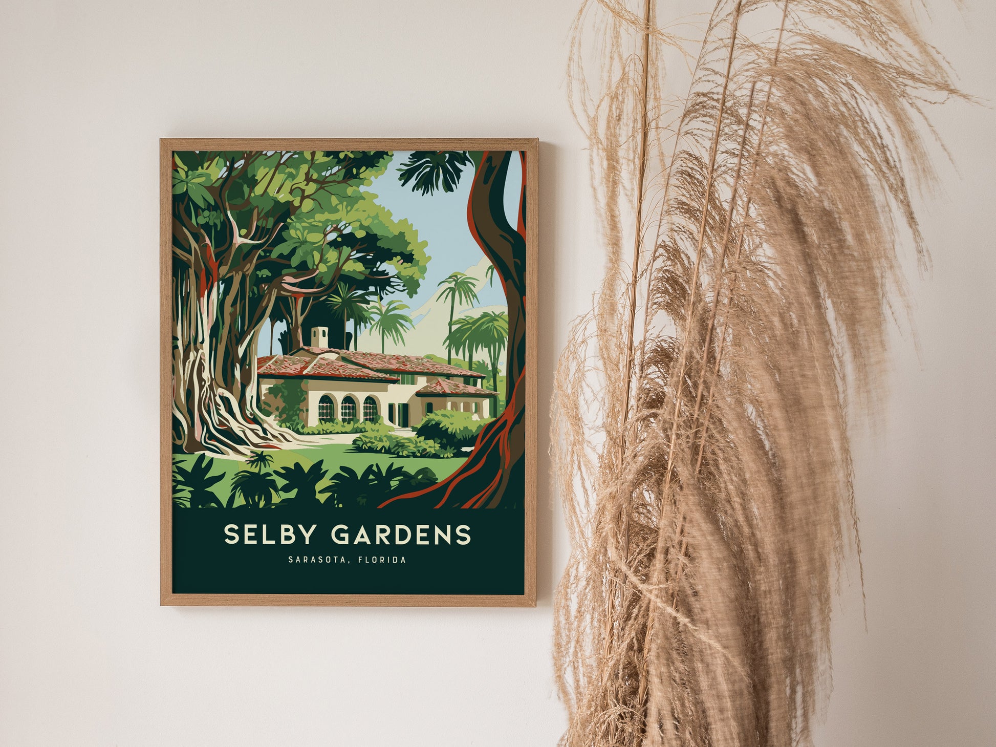 A framed poster titled Selby Gardens showcases lush tropical trees and a building, set against a white wall beside dried pampas grass.