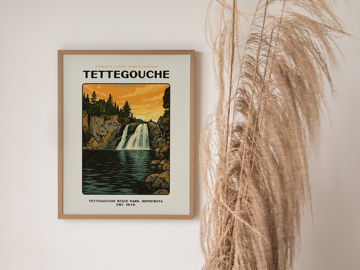 Framed Tettegouche State Park poster depicting a waterfall surrounded by cliffs and trees against a vibrant sunset. Dried grasses are displayed beside the artwork on a white wall.