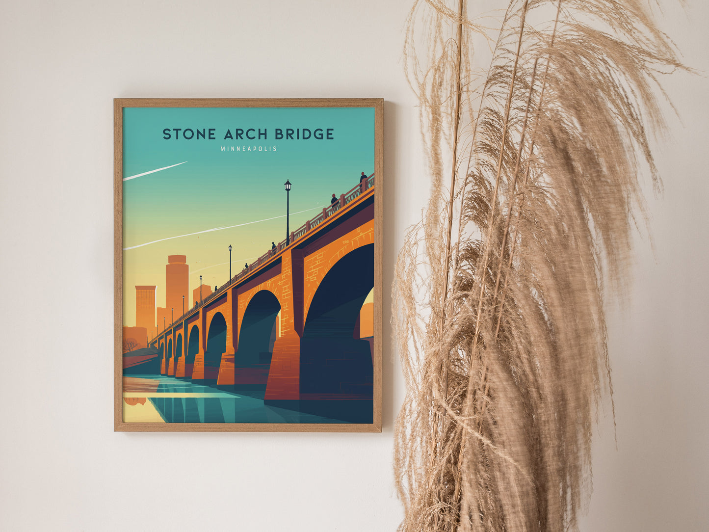 A framed poster of the Stone Arch Bridge in Minneapolis is displayed on a wall next to tall, wispy dried grasses. The artwork features a vibrant sunset skyline.