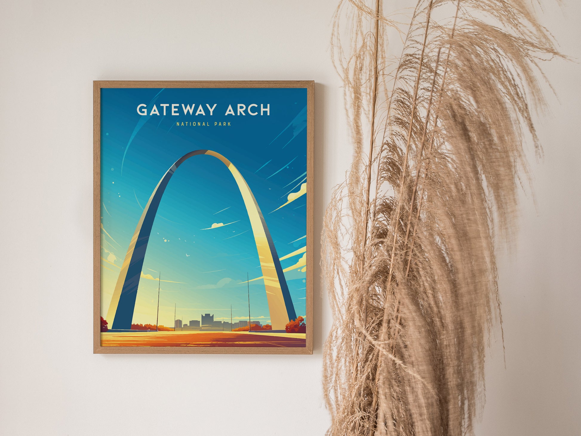 A framed poster of the Gateway Arch in St. Louis, set against a blue sky, hangs on a wall beside dried pampas grass.