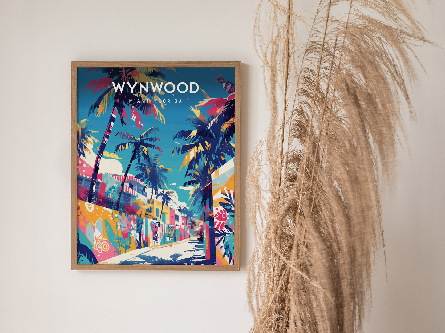 A vibrant poster of Wynwood, Miami, featuring colorful street art and towering palm trees, is framed and displayed on a white wall. Dried plants are positioned to the right, adding a natural element to the scene.