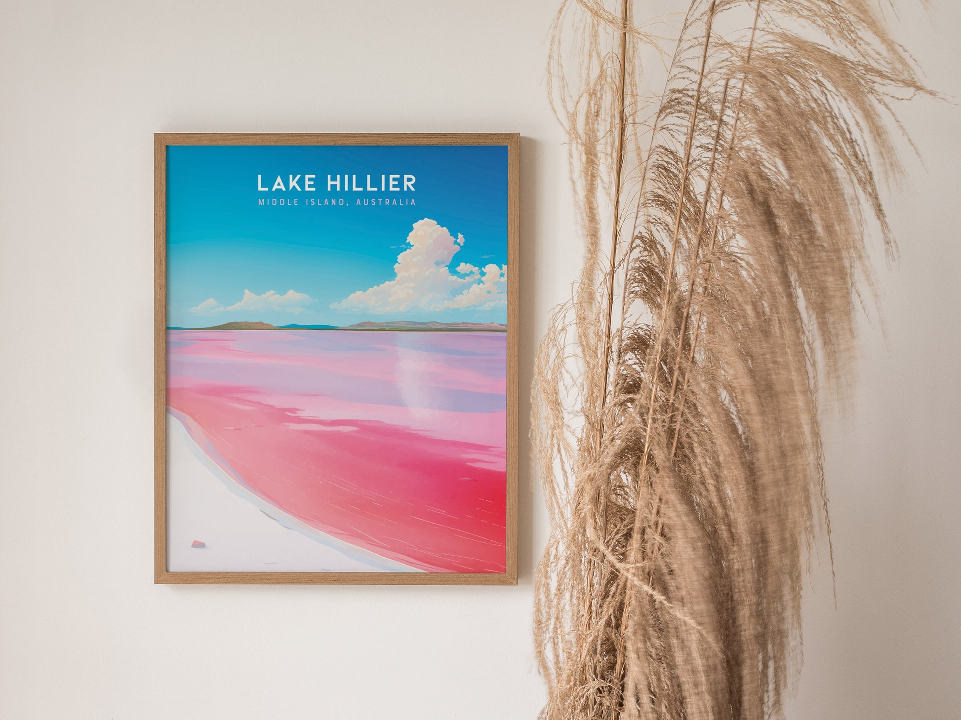 Framed poster of Lake Hillier features vibrant pink water contrasting with blue sky, displayed on a white wall next to tall, dried grasses.