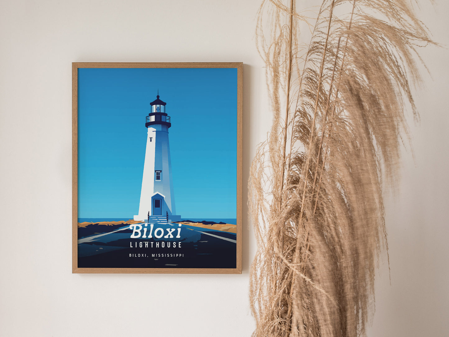 A framed Biloxi Lighthouse poster hangs on a wall next to dried pampas grass. The artwork features a minimalistic depiction of the lighthouse against a deep blue sky.
