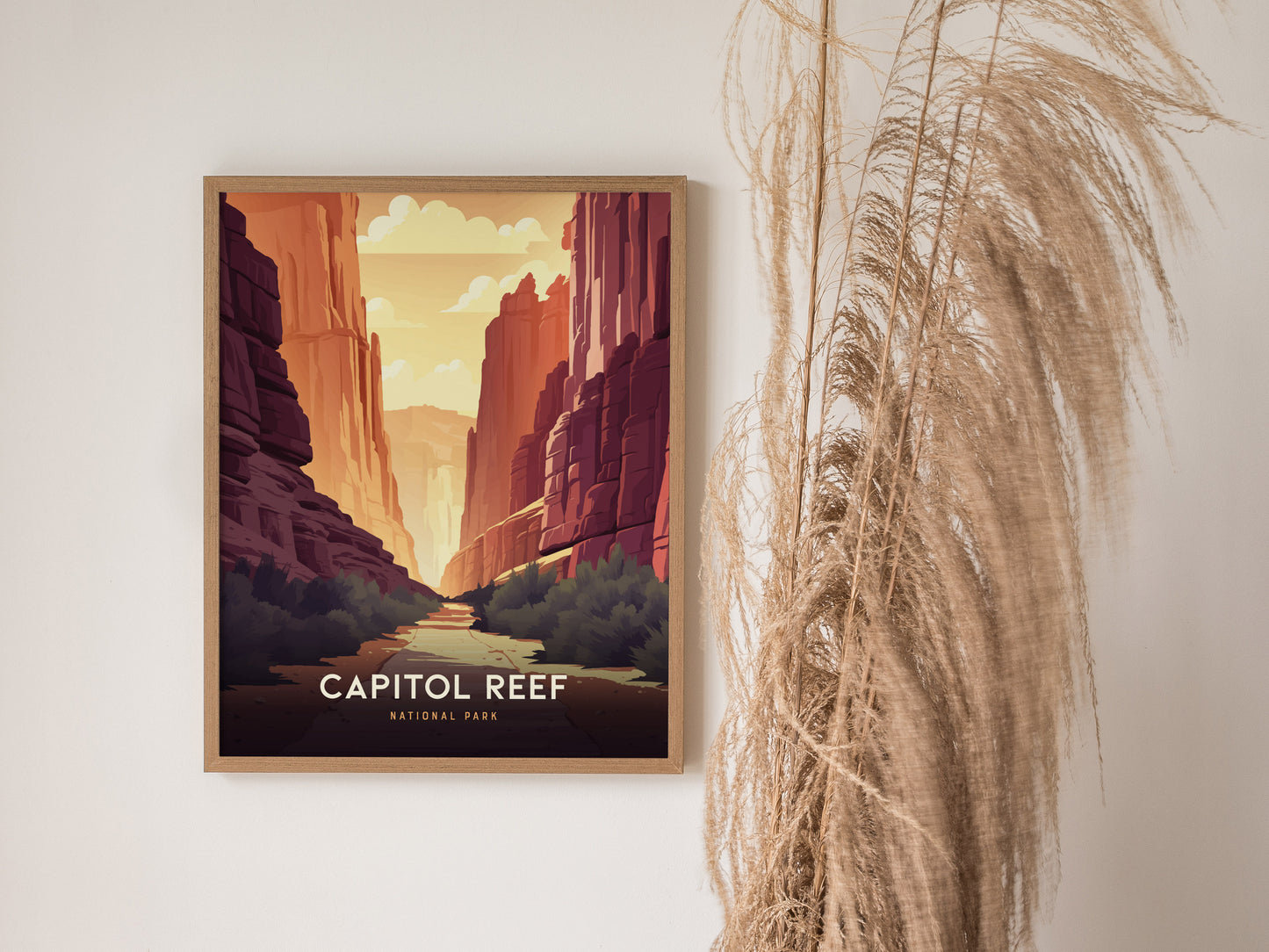 Framed artwork depicting a canyon landscape with towering red cliffs and a sunny sky, labeled Capitol Reef National Park. Set against a neutral wall, accompanied by dried pampas grass.