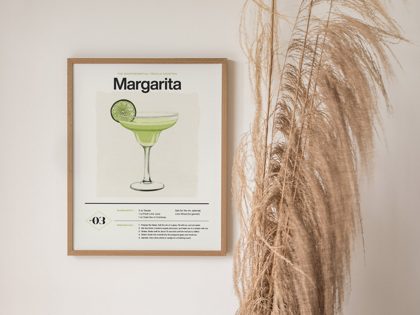 Framed Margarita cocktail poster featuring an illustration of a drink garnished with a lime slice, hanging on a white wall next to tall, dried pampas grass.