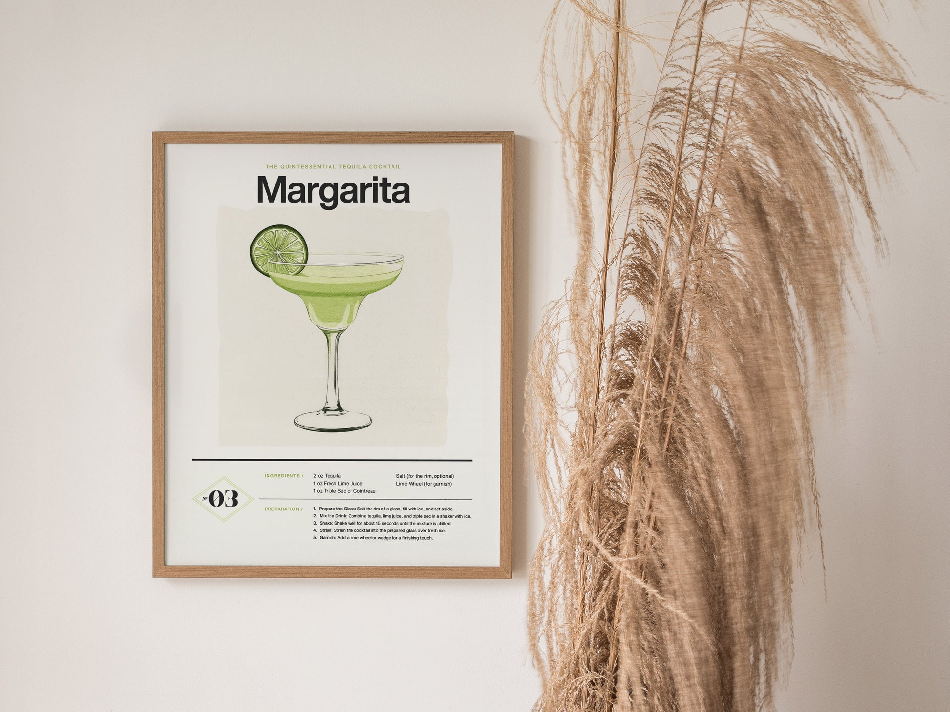 Framed Margarita cocktail poster featuring an illustration of a drink garnished with a lime slice, hanging on a white wall next to tall, dried pampas grass.