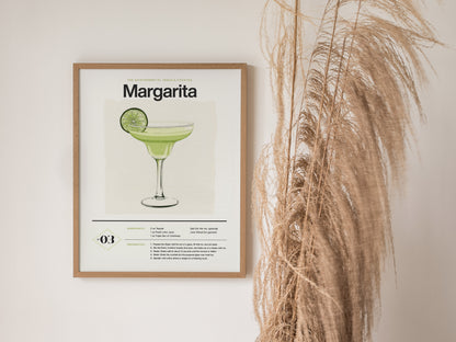 Framed Margarita cocktail poster featuring an illustration of a drink garnished with a lime slice, hanging on a white wall next to tall, dried pampas grass.