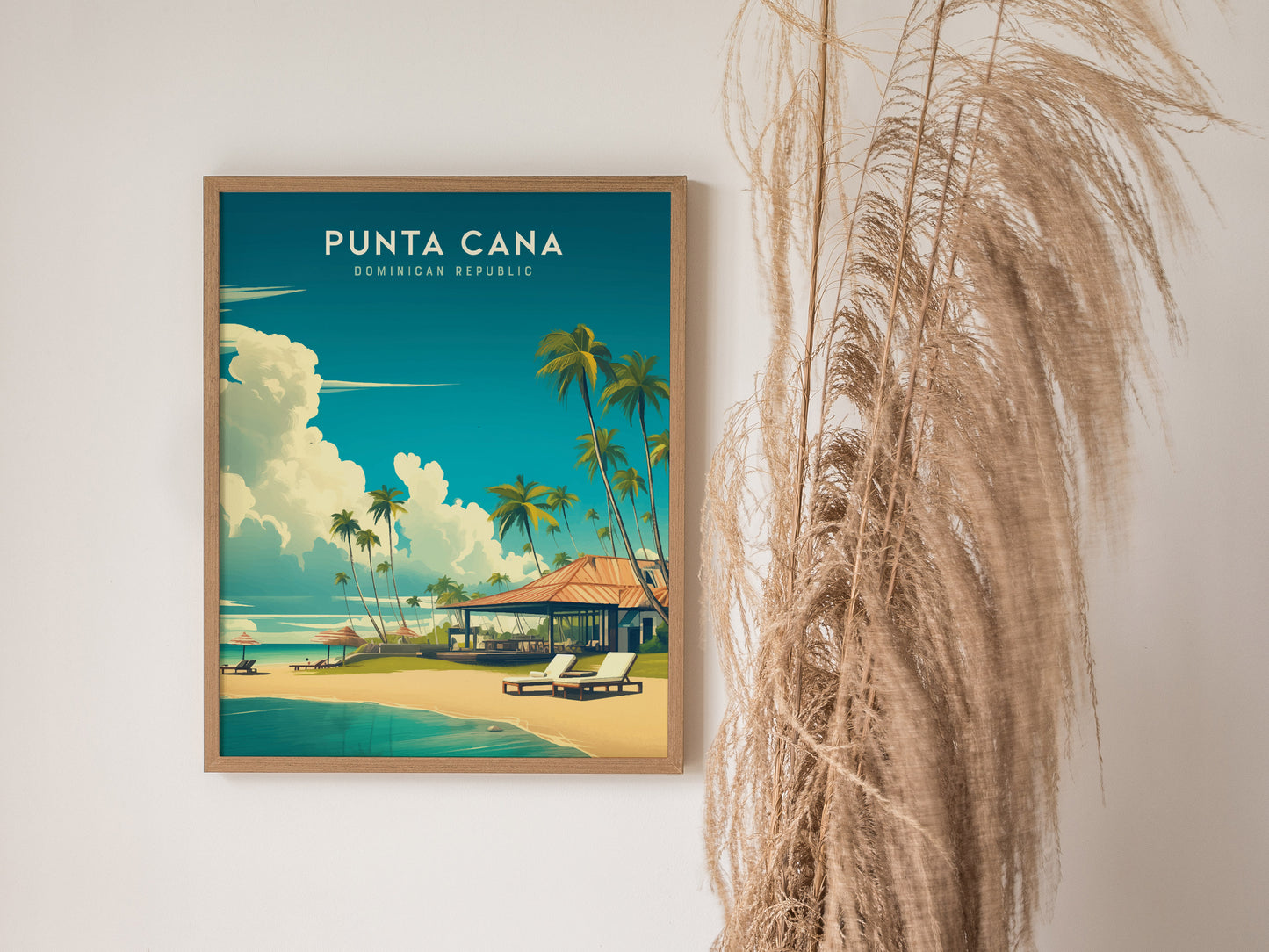 A framed poster of Punta Cana, Dominican Republic, featuring a tropical beach scene with palm trees, clear blue skies, and white sand. The artwork is displayed on a white wall next to tall, dry pampas grass.