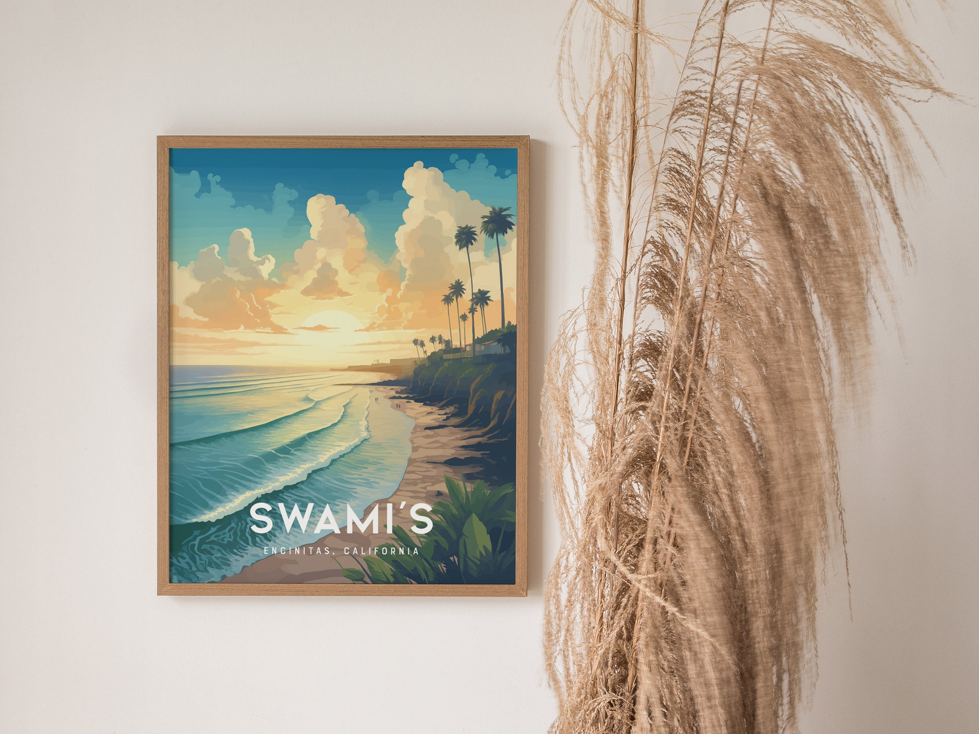 Framed artwork depicts a sunset over Swamis Beach in Encinitas, California, with waves and palm trees. Tall dried grasses are positioned beside the poster on a white wall.